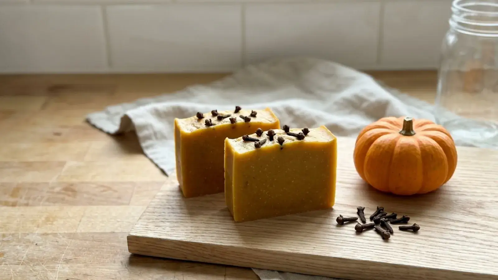 Handmade yellow soap bars topped with cloves sit beside a small pumpkin on a wooden cutting board.