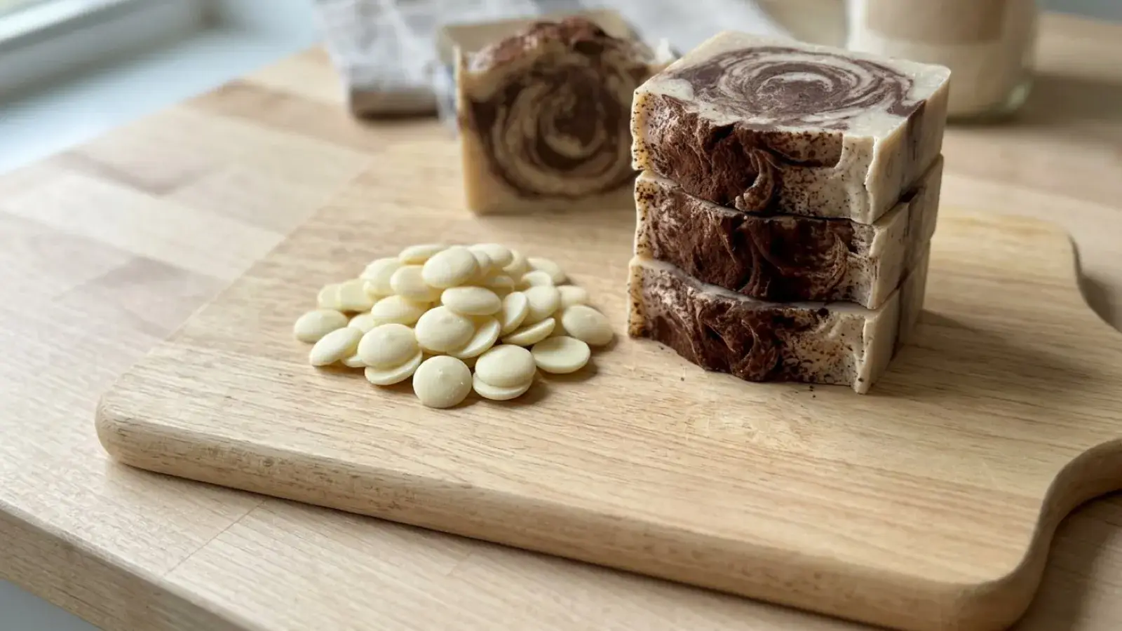 Stacked brown and cream swirled soap bars beside cocoa butter wafers on a wooden cutting board.