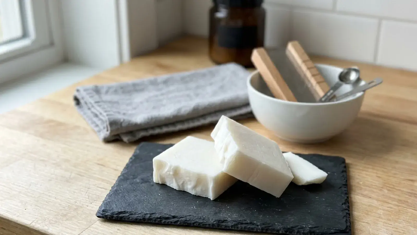 Bars of white handmade soap rest on a slate board beside soap-making tools on a wooden countertop.