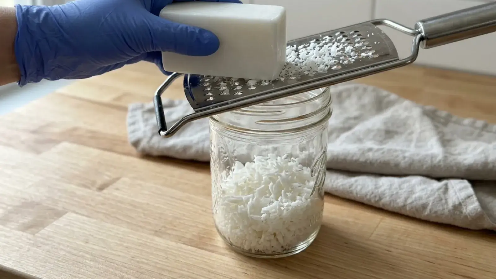 A gloved hand grates a white bar of soap into a glass jar using a metal grater.